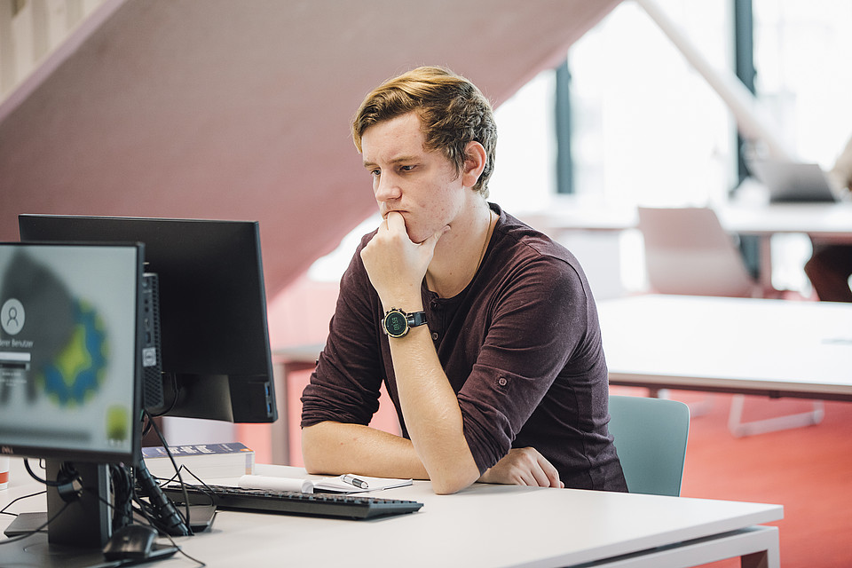 Studierender schaut in einem Lernraum der Universitätsbibliothek mit konzentriertem Blick auf einen Computerbildschirm.