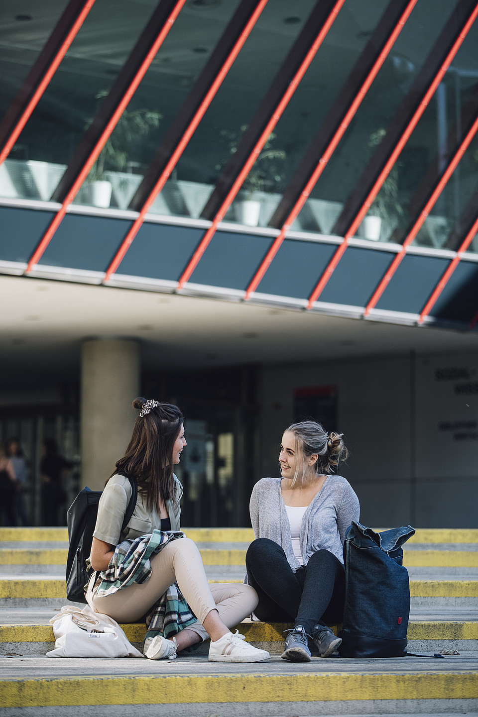 Zwei Studierende sitzen auf den Stufen vor einem Gebäude am Campus und unterhalten sich