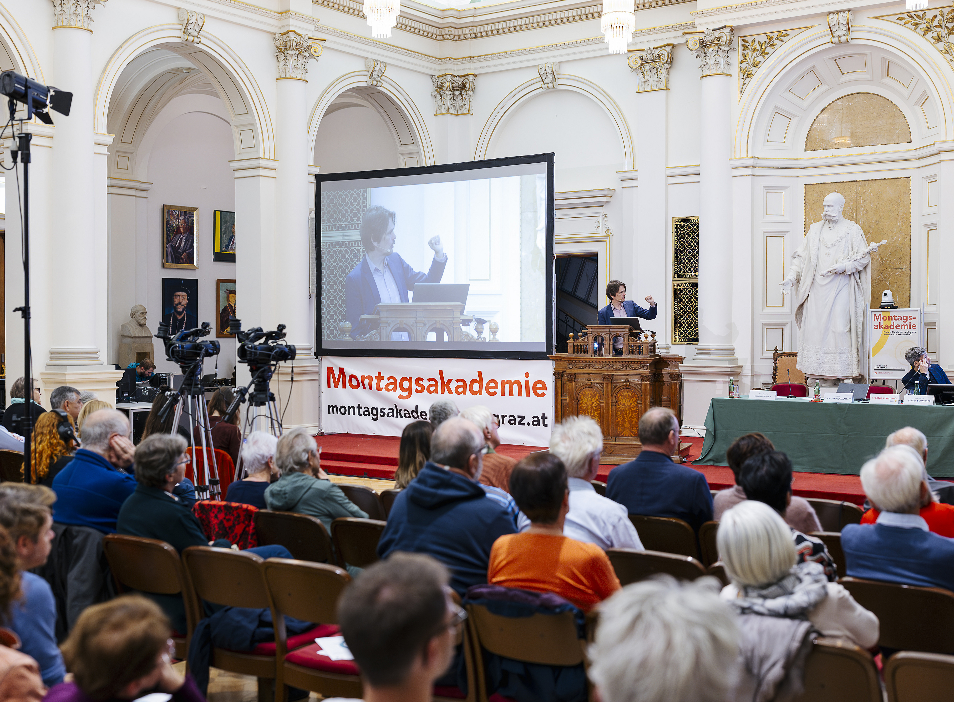 A lecture is taking place in a grand hall with tall white columns and golden ornamentation. In the foreground, numerous audience members are seated on chairs. In the background, a speaker stands at a wooden lectern; her gestures are projected onto a large screen. Several cameras are set up on the left; on the right, another person is seated at a table with a microphone. A large statue stands in the hall, and a banner reading “Monday Academy” hangs on the wall. ©KANIZAJ photography 2024