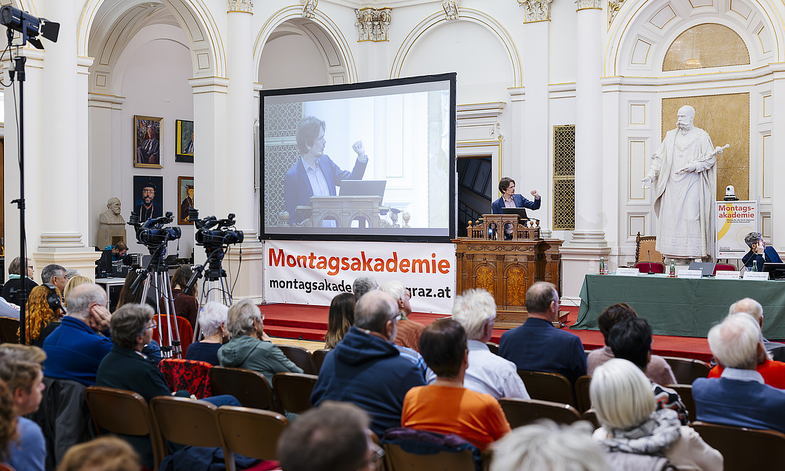 A lecture is taking place in a grand hall with tall white columns and golden ornamentation. In the foreground, numerous audience members are seated on chairs. In the background, a speaker stands at a wooden lectern; her gestures are projected onto a large screen. Several cameras are set up on the left; on the right, another person is seated at a table with a microphone. A large statue stands in the hall, and a banner reading “Monday Academy” hangs on the wall. 