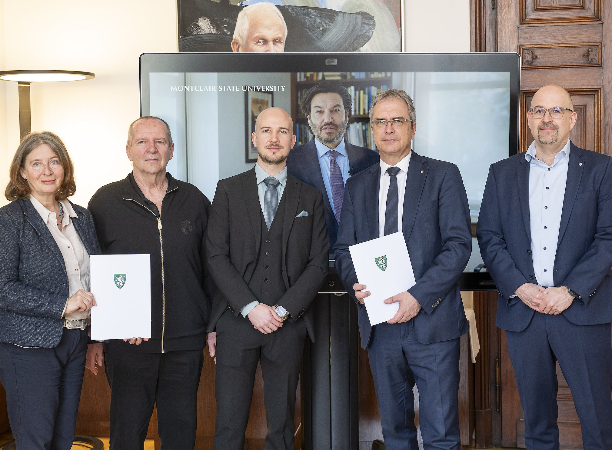 In einem holzvertäfelten Besprechungsraum stehen sechs Personen in einer Reihe und blicken in Richtung Kamera. Die meisten tragen dunkle Anzüge oder Businesskleidung, eine Person ist leger in einer dunklen Jacke gekleidet. Zwei Personen halten weiße Mappen mit einem grünen Wappen auf der Vorderseite. Hinter der Gruppe ist ein großer Bildschirm zu sehen, auf dem der Schriftzug „Montclair State University“ sowie zwei weitere Personen in einem Videocall erscheinen. Rechts im Hintergrund befindet sich eine schwere Holztür, links eine Stehleuchte. ©Foto Fischer, Graz