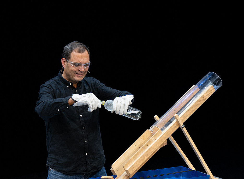Eine Person trägt weiße Handschuhe und hält eine Plastikflasche über einem blauen Behälter mit einem schräg montierten transparenten Rohr auf einer Holzhalterung. 