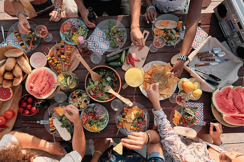 View from above of bowls with various dishes on a wooden table and arms of people taking something for themselves