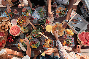 View from above of bowls with various dishes on a wooden table and arms of people taking something for themselves