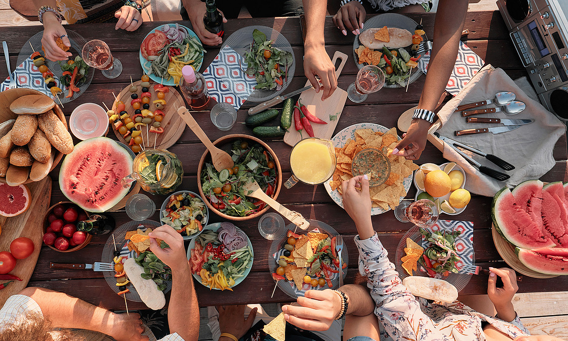 View from above of bowls with various dishes on a wooden table and arms of people taking something for themselves