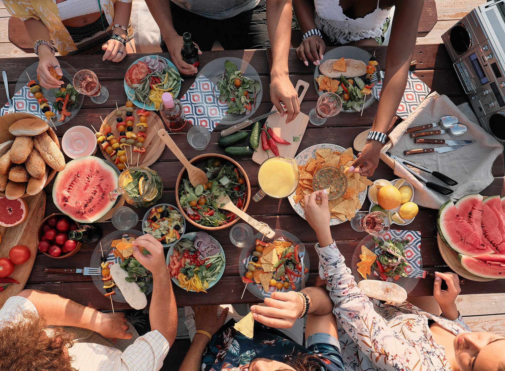 View from above of bowls with various dishes on a wooden table and arms of people taking something for themselves ©AnnaStills - stock.adobe.com 