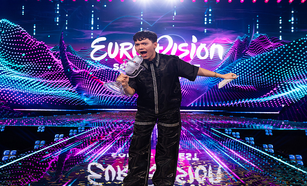 Ein junger Mann steht mit einer Trophäe in der Hand auf einer Bühne. Hinter seinem Kopf leuchtet der Titel "Eurovision" auf einer LED-Leinwand.