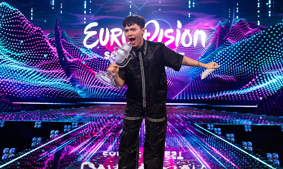 Ein junger Mann steht mit einer Trophäe in der Hand auf einer Bühne. Hinter seinem Kopf leuchtet der Titel "Eurovision" auf einer LED-Leinwand.