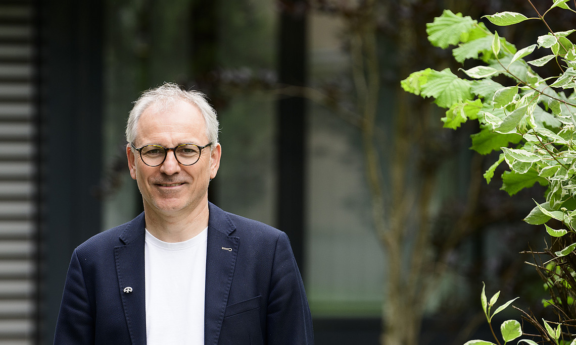 Älterer Mann mit Brille in dunkelblauem Blazer und weißem T‑Shirt steht draußen vor einem Gebäude, rechts im Bild grüne Sträucher.