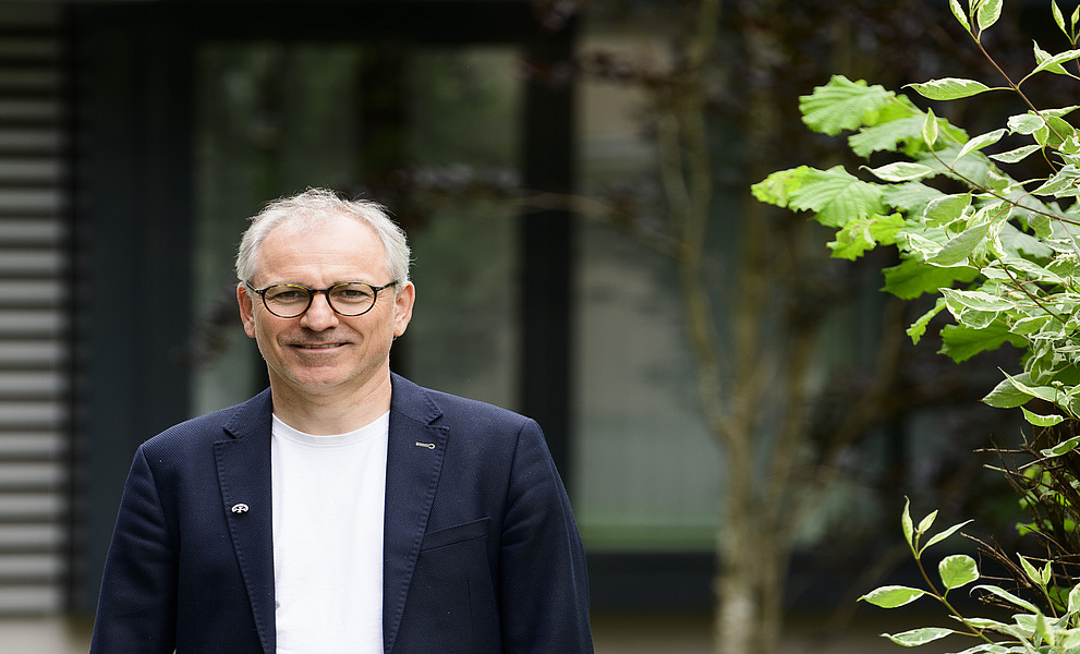 Älterer Mann mit Brille in dunkelblauem Blazer und weißem T‑Shirt steht draußen vor einem Gebäude, rechts im Bild grüne Sträucher.