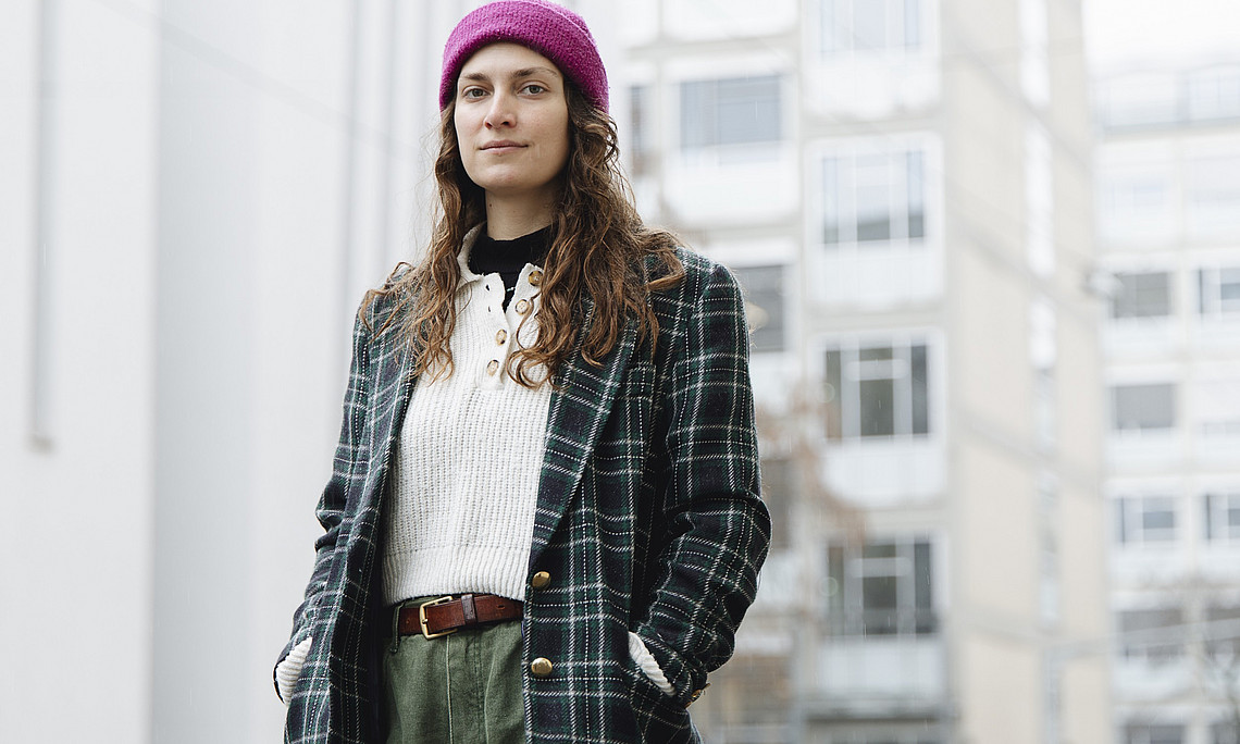  A young adult is standing outdoors, slightly to the left in the foreground, with their upper body facing the camera. Long curly hair falls onto their shoulders; they are wearing a purple knitted hat. The person is wearing a dark green checked coat with gold buttons, underneath a white knitted jumper with a button placket, a brown leather belt and olive-green trousers; both hands are in the coat pockets. In the background are several tall, light-coloured residential or office buildings.
