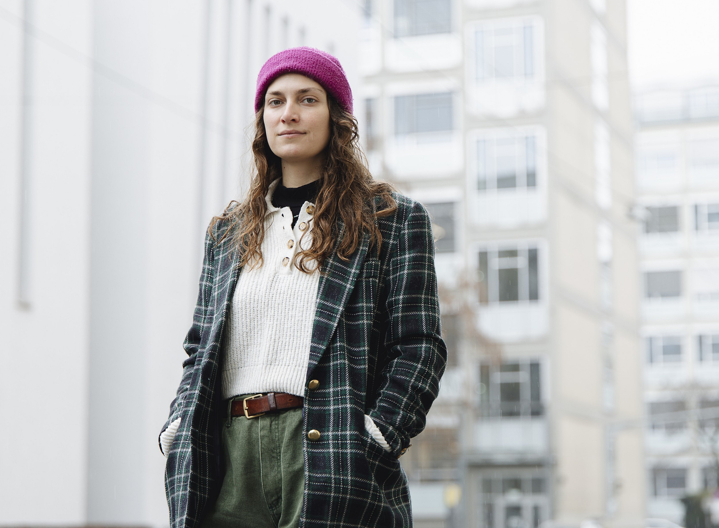  A young adult is standing outdoors, slightly to the left in the foreground, with their upper body facing the camera. Long curly hair falls onto their shoulders; they are wearing a purple knitted hat. The person is wearing a dark green checked coat with gold buttons, underneath a white knitted jumper with a button placket, a brown leather belt and olive-green trousers; both hands are in the coat pockets. In the background are several tall, light-coloured residential or office buildings. ©KANIZAJ 2022
