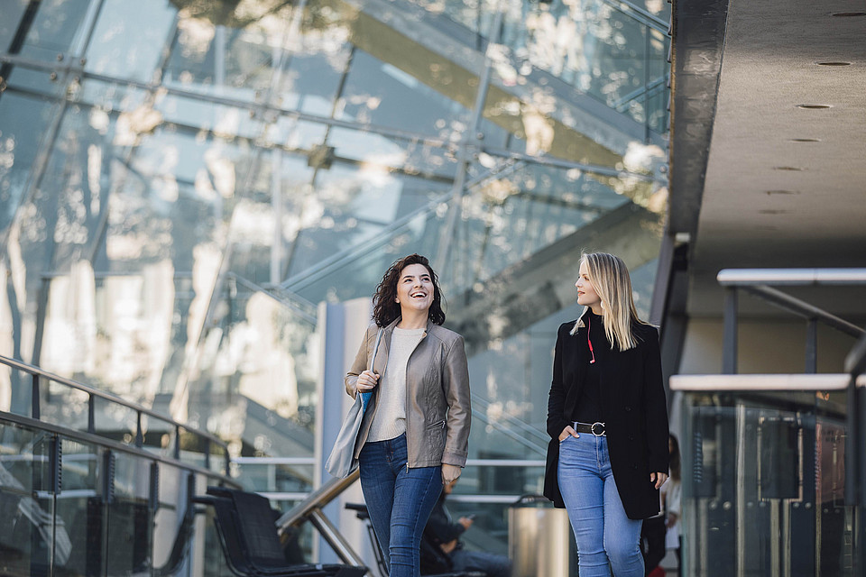 Zwei Studierende flanieren über den Campus, hinter ihnen liegt ein modernes verglastes Gebäude, Studierenden vor dem Hauptgebäude der Uni Graz, Sujetbild fürs Studium Uni Graz