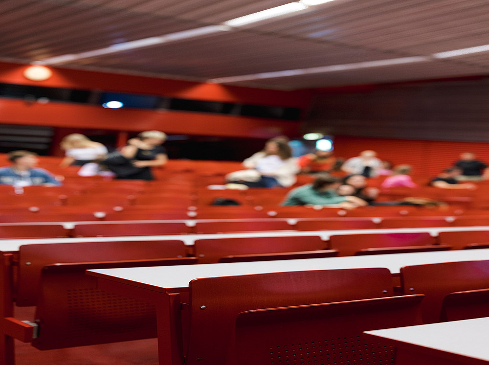 Auf dem Bild ist ein Hörsaal mit roten Stühlen zu sehen, während die Studierenden im Hintergrund verschwommen dargestellt sind. ©Uni Graz/Kanizaj