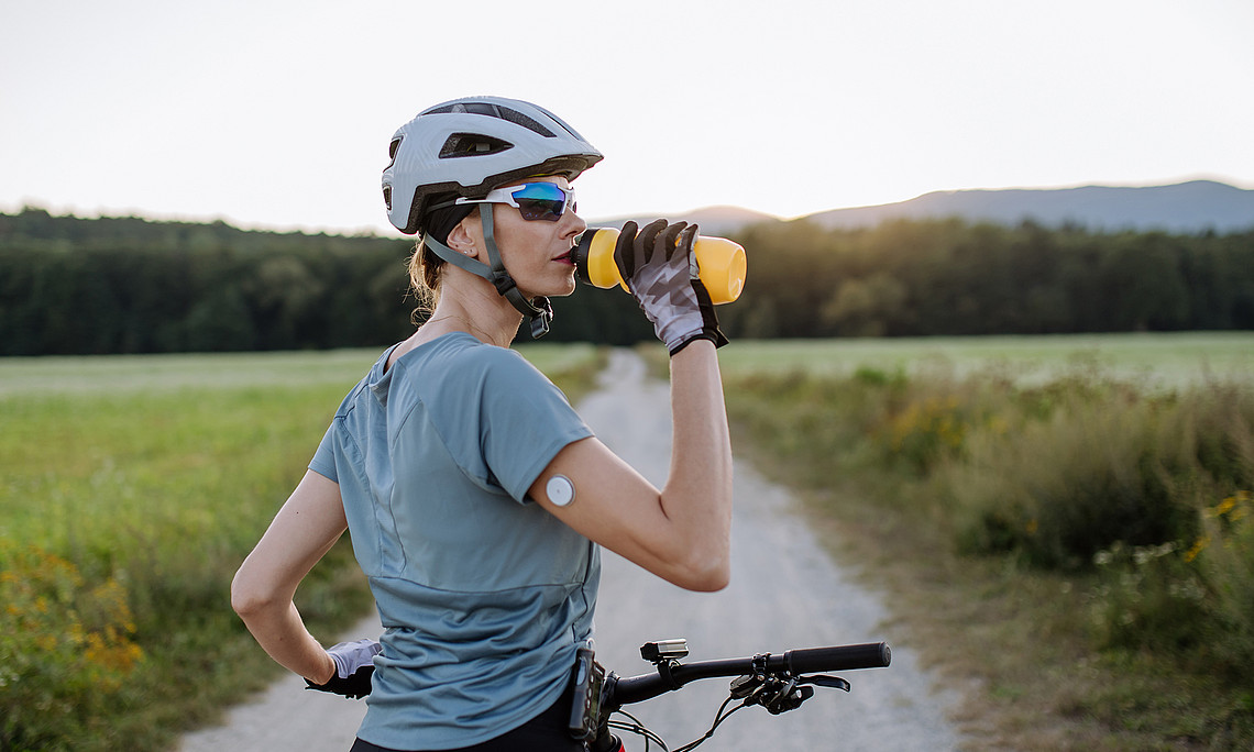 Radfahrerin mit Diabetes und Glukose-Monitor am Arm, die während einer Tour Wasser trinkt