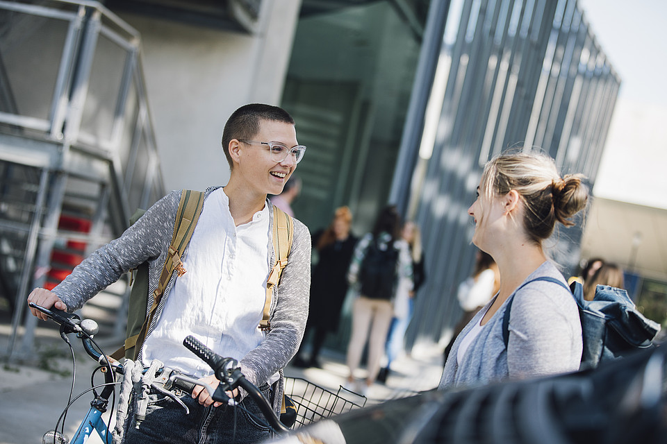 Im Vordergrund unterhalten sich zwei Studierende, während sie ihre Fahrräder halten; dahinter ist vor einem modernen verglasten Gebäude eine weitere Studierendengruppe zu sehen 