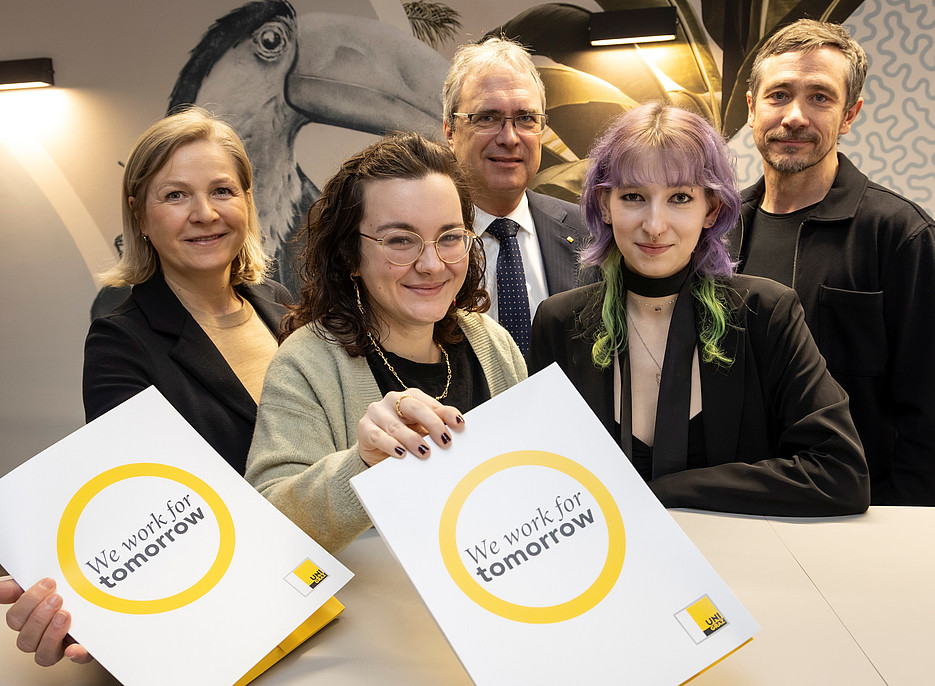 Eine Gruppe von Personen steht um einen Tisch. Zwei von ihnen halten weiße Mappen mit einem gelben Kreis und der Aufschrift „We work for tomorrow“. Im Hintergrund ist eine Wand mit einem großflächigen Vogelmotiv und Pflanzenillustrationen zu sehen, beleuchtet von modernen Wandlampen. 