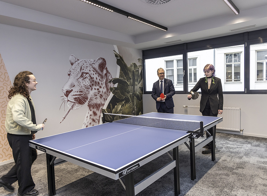 Drei Personen stehen um einen Tischtennistisch in einem modern gestalteten Raum. Eine Person spielt aktiv, während die anderen zuschauen und Tischtennisschläger halten. Die Wand im Hintergrund ist mit einem großen Leopardenmotiv und Pflanzenillustrationen dekoriert. Große Fenster lassen Tageslicht in den Raum. ©Helmut Lunghammer