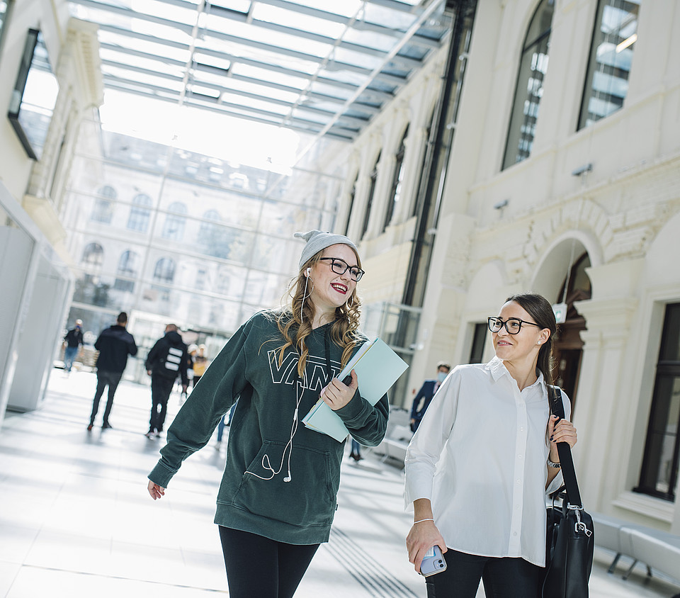 Zwei Studierende im Vordergrund unterhalten sich während sie im hellen Eingangsbereich der Universitätsbibliothek gehen, im Hintergrund bewegen sich Studierende weg