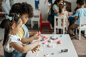 Zwei Kinder spielen mit Plastilin an einem Tisch, im Hintergrund ist eine weitere Kindergruppe zu sehen, die auch an einem Tisch sitzt.