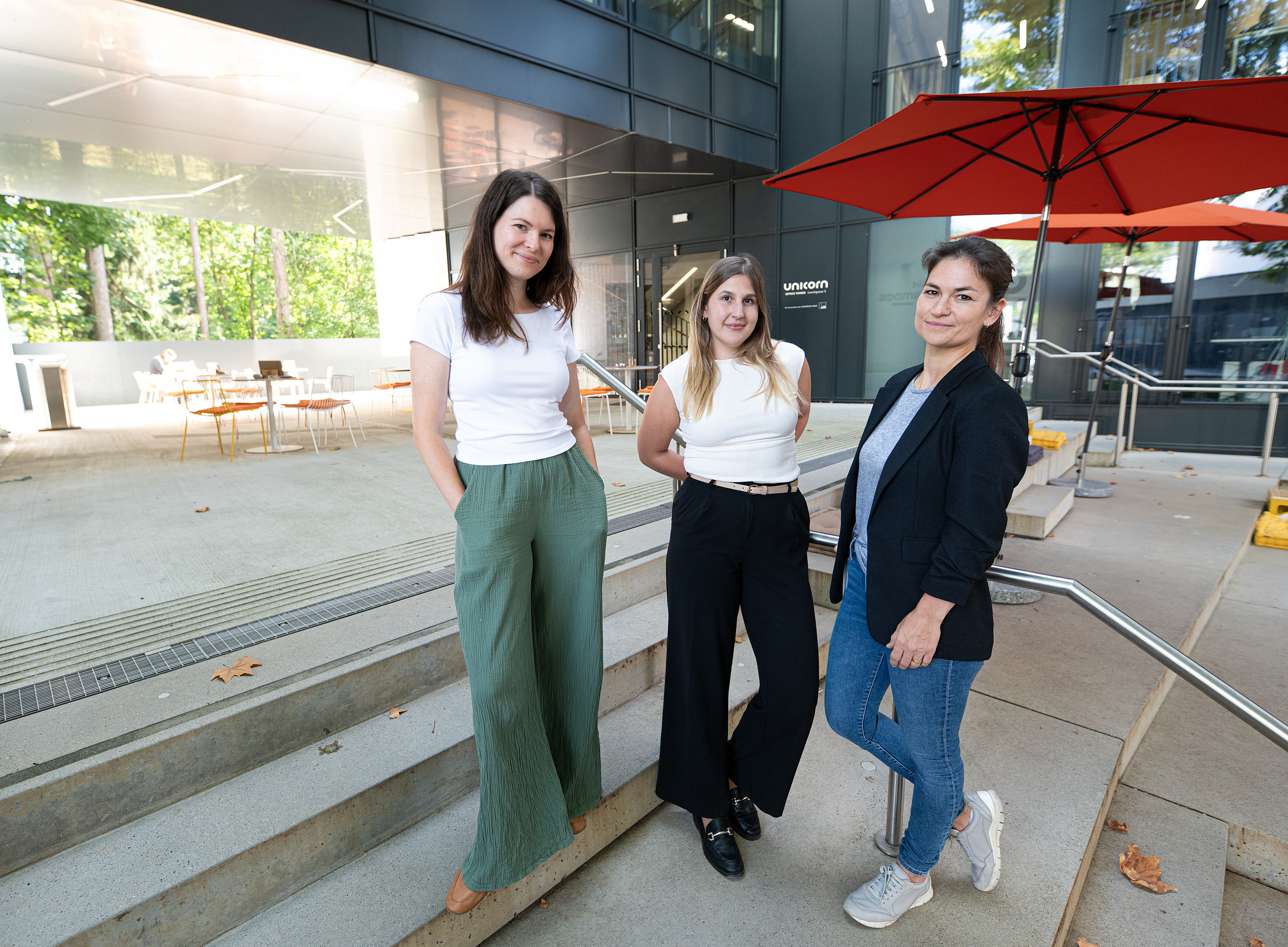 Drei junge Frauen in eleganter Geschäftskleidung stehen auf den Stufen einer Terrasse mit Sitzgelegenheiten und Sonnenschirmen. ©Uni Graz/Tzivanopoulos