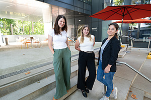 three young women in stylish business attire stand on the steps of an outdoor patio with seating and umbrellas. 