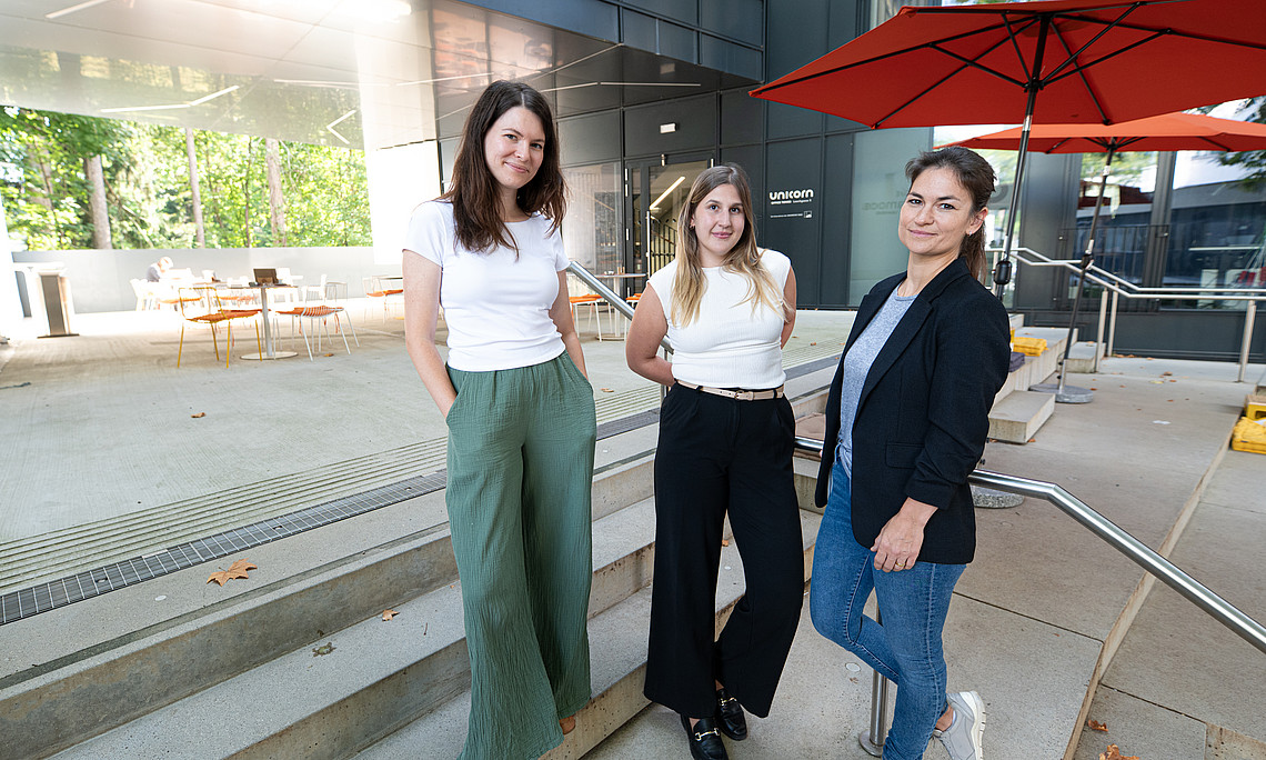 three young women in stylish business attire stand on the steps of an outdoor patio with seating and umbrellas. 
