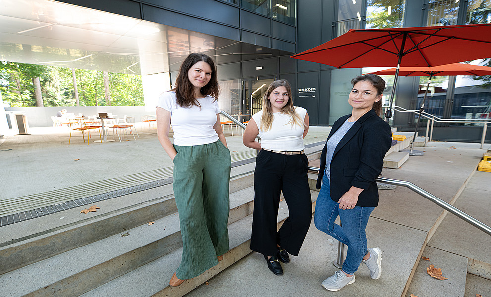 Drei junge Frauen in eleganter Geschäftskleidung stehen auf den Stufen einer Terrasse mit Sitzgelegenheiten und Sonnenschirmen.