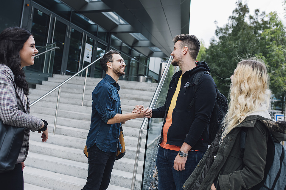 Vier Studierende unterhalten sich vor dem Chemie-Gebäude miteinander; im Mittelpunkt stehen zwei Personen, die sich die Hände reichen und einander anlächeln