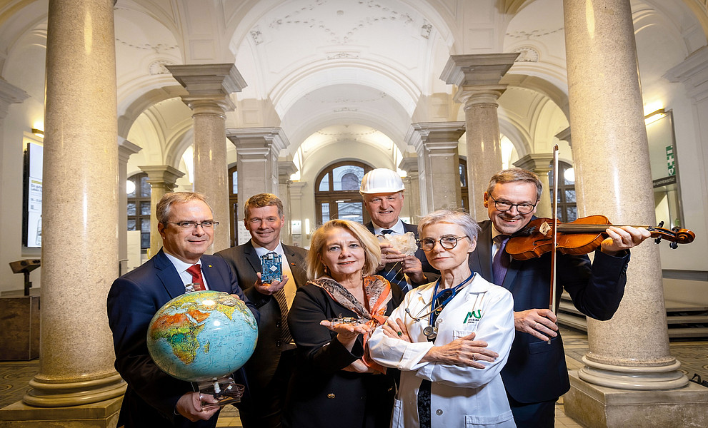 Gruppe von sechs Personen in festlicher Kleidung steht in einem historischen Foyer mit hohen Gewölbedecken und Marmorsäulen. Sie halten symbolische Objekte aus verschiedenen Fachbereichen in den Händen – unter anderem einen Globus, ein elektronisches Bauteil, ein Architekturmodell, eine Muschel, eine Violine sowie ein Stethoskop – und repräsentieren damit die Vielfalt von Wissenschaft, Medizin, Technik und Kunst.
