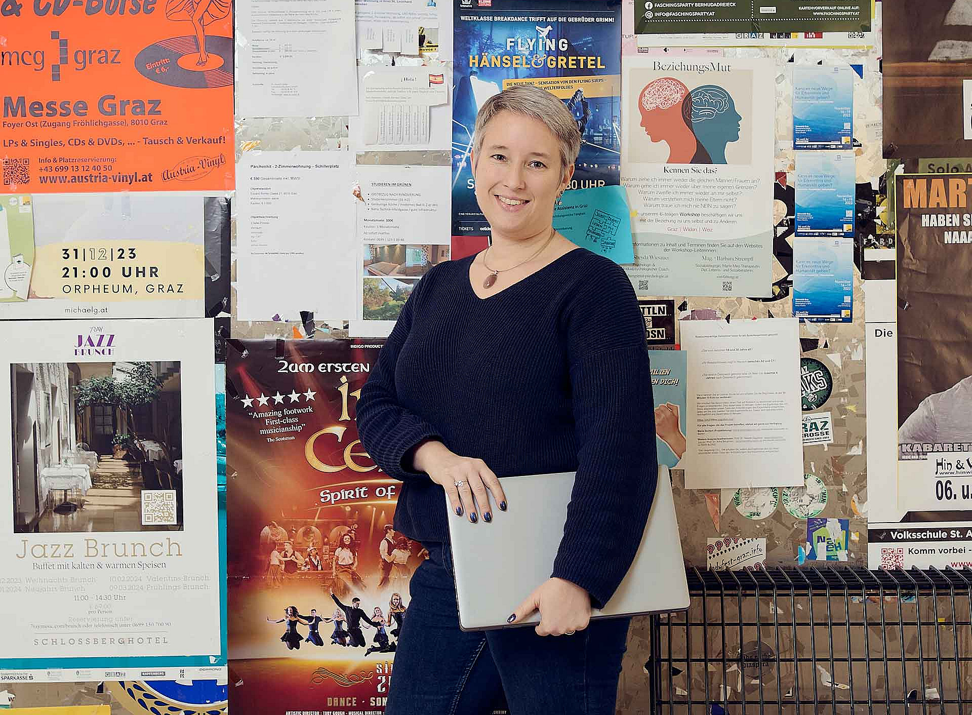 A short-haired woman is standing, smiling, in front of a noticeboard covered in colourful posters, holding a closed laptop in their hand. 