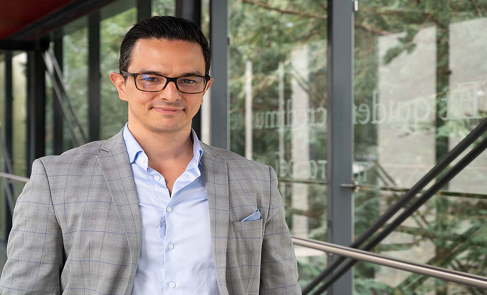 Porträt einer Person mit Brille in hellgrauem Sakko und hellblauem Hemd in einem modernen Bürogebäude mit großen Fensterfronten und Blick ins Grüne.