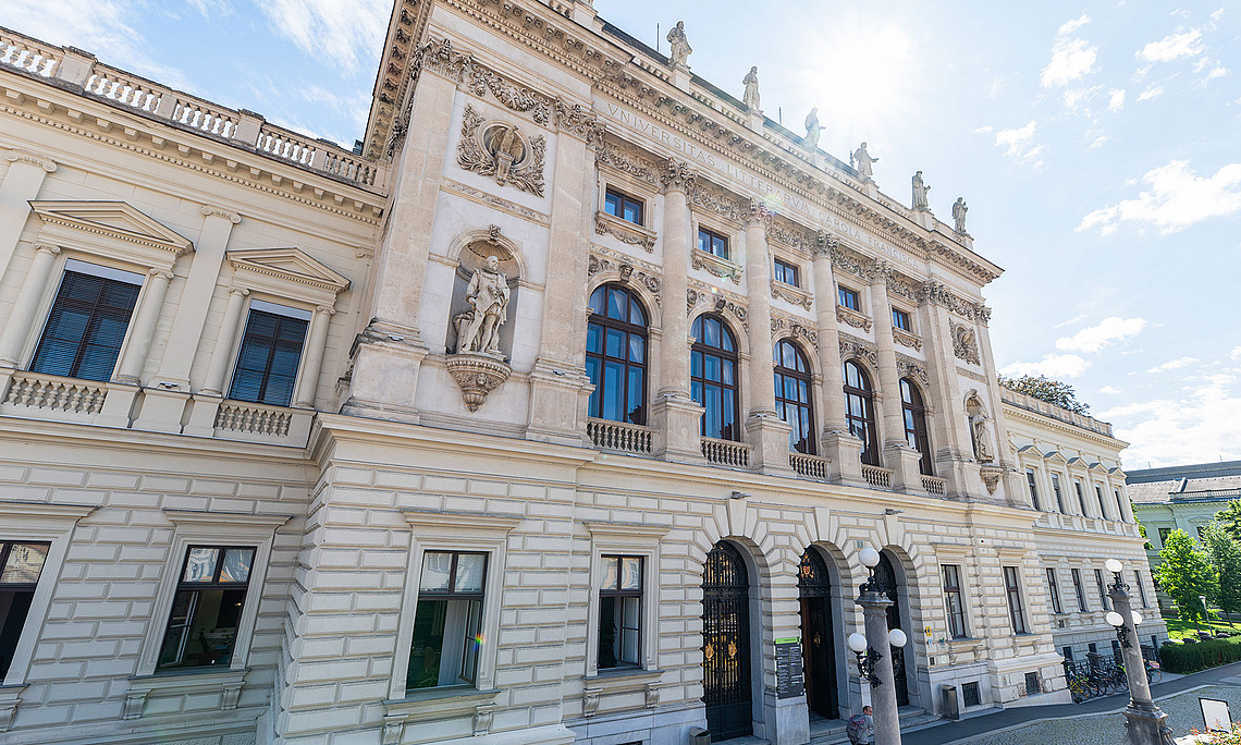 Monumentale neobarocke Fassade des Hauptgebäudes der Universität Graz mit hellen Natursteinquadern, hohen Rundbogenfenstern, kräftigen Säulen und Nischenfiguren, im leicht schrägen Straßenblick bei hellem Tageslicht
