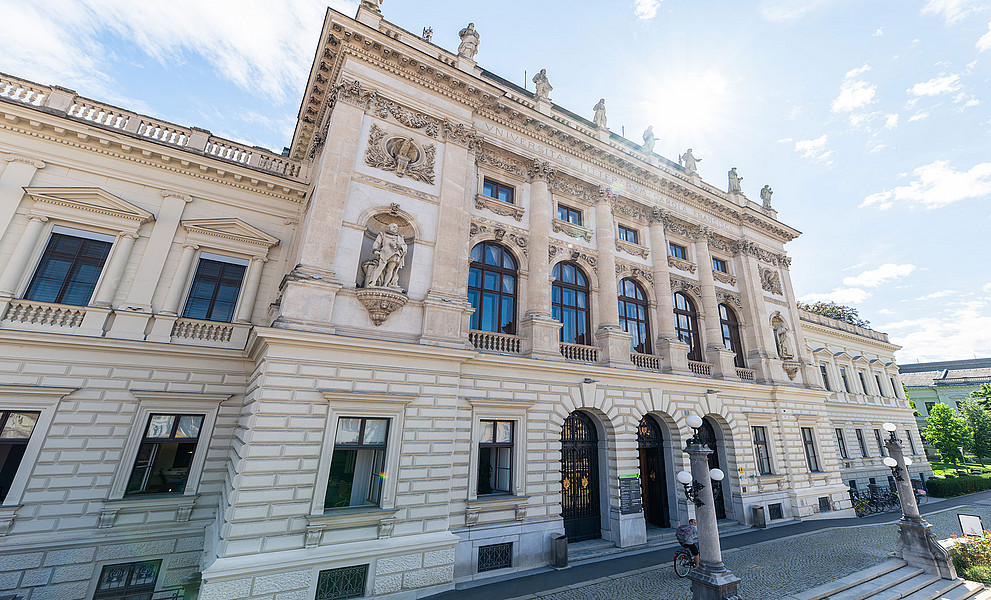 Monumentale neobarocke Fassade des Hauptgebäudes der Universität Graz mit hellen Natursteinquadern, hohen Rundbogenfenstern, kräftigen Säulen und Nischenfiguren, im leicht schrägen Straßenblick bei hellem Tageslicht