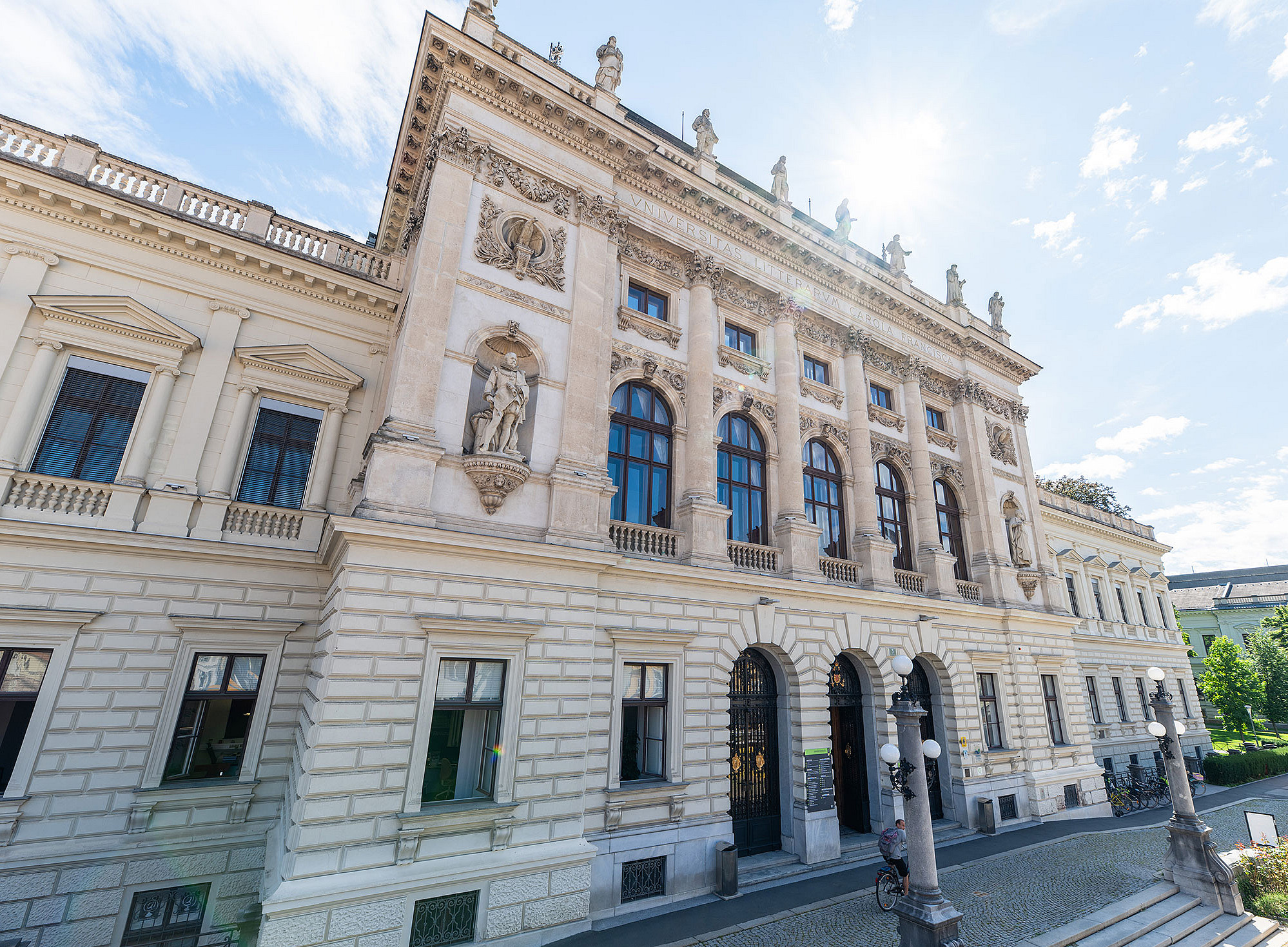 Monumentale neobarocke Fassade des Hauptgebäudes der Universität Graz mit hellen Natursteinquadern, hohen Rundbogenfenstern, kräftigen Säulen und Nischenfiguren, im leicht schrägen Straßenblick bei hellem Tageslicht ©David Schreyer