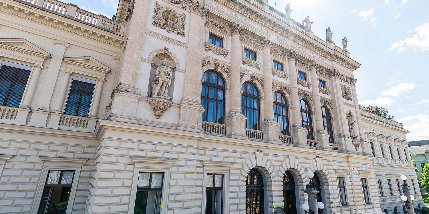 Monumentale neobarocke Fassade des Hauptgebäudes der Universität Graz mit hellen Natursteinquadern, hohen Rundbogenfenstern, kräftigen Säulen und Nischenfiguren, im leicht schrägen Straßenblick bei hellem Tageslicht
