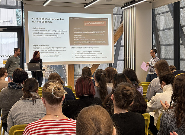 zwei Frauen und ein Mann stehen vor einem Tisch mit Laptop und halten einen Vortrag, hinter ihnen sieht man die Leinwand mit der Präsentation. Im Publikum sitzen viele Menschen und hören zu ©Uni Graz/Schmieder