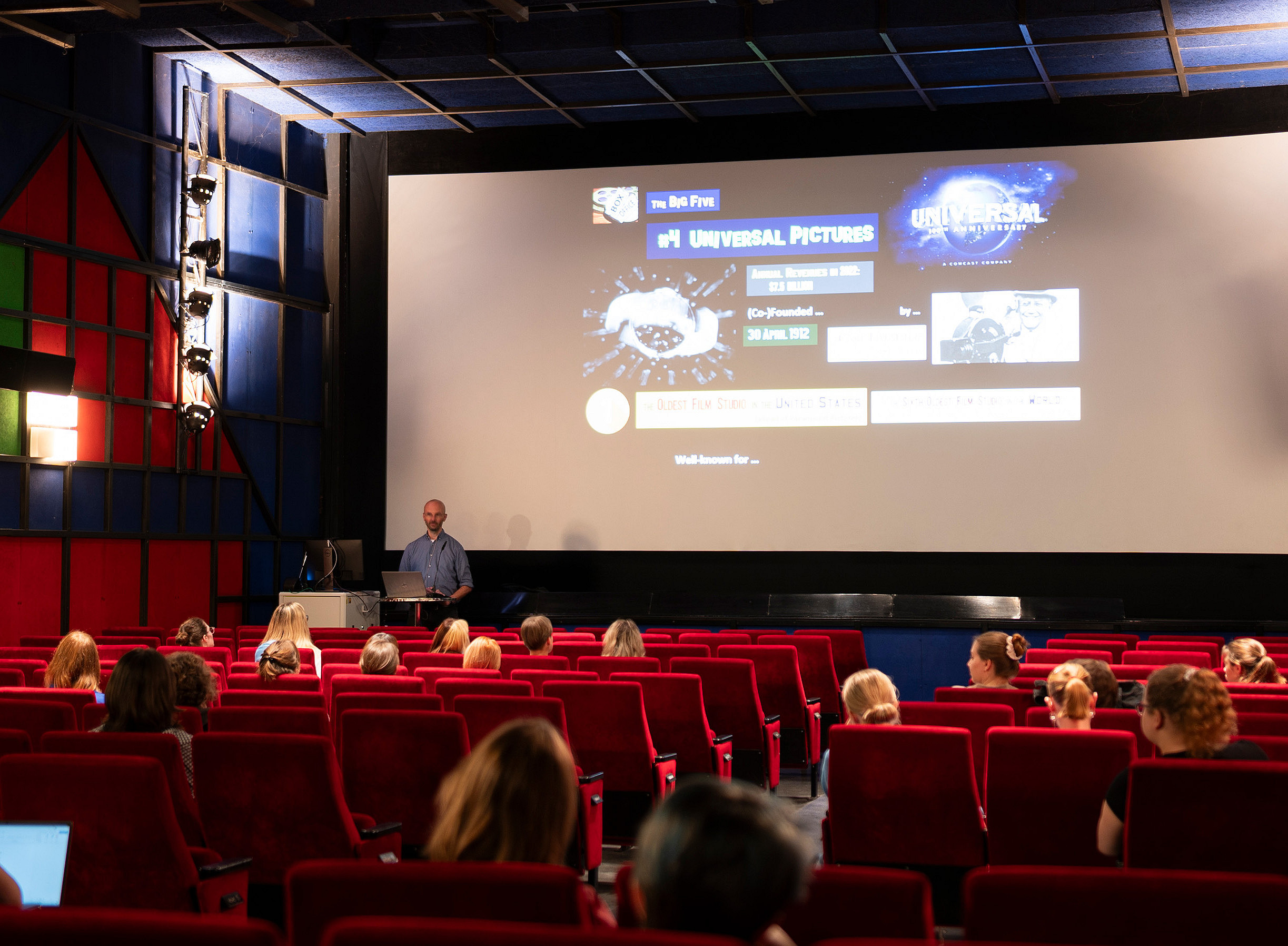 Ein Kinosaal mit einigen besetzten Plätzen und einem Mann vor der Leinwand, die einen Vortrag hält. ©Uni Graz/Angele
