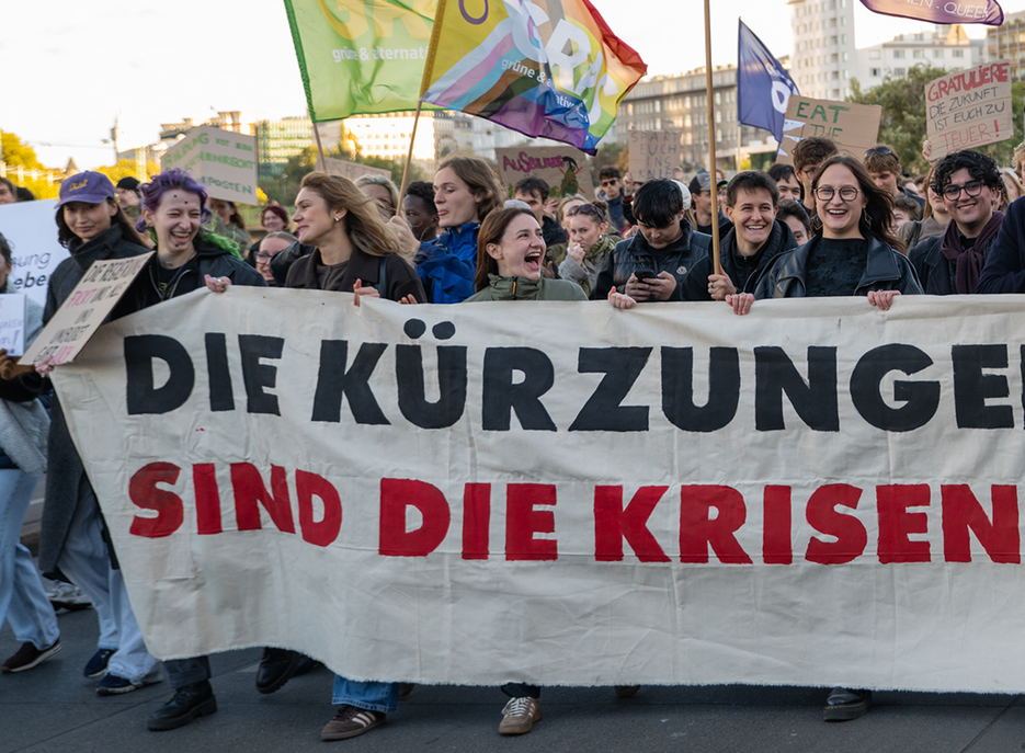 Demonstrierende tragen ein großes Banner mit der Aufschrift „DIE KÜRZUNGEN SIND DIE KRISEN“, darüber wehen Regenbogen- und Parteifahnen. 