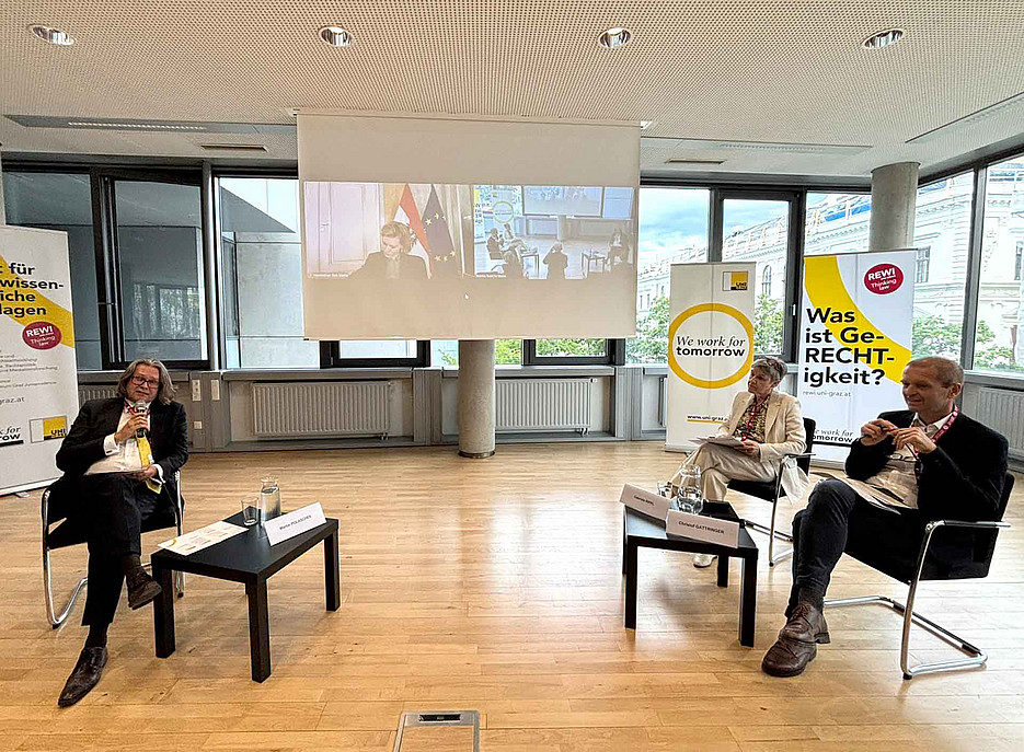 Drei Erwachsene sitzen in einem hellen Konferenzraum auf Stühlen, diskutieren, vor ihnen kleine Tische; im Hintergrund Banner und eine Leinwand mit Videokonferenz. 