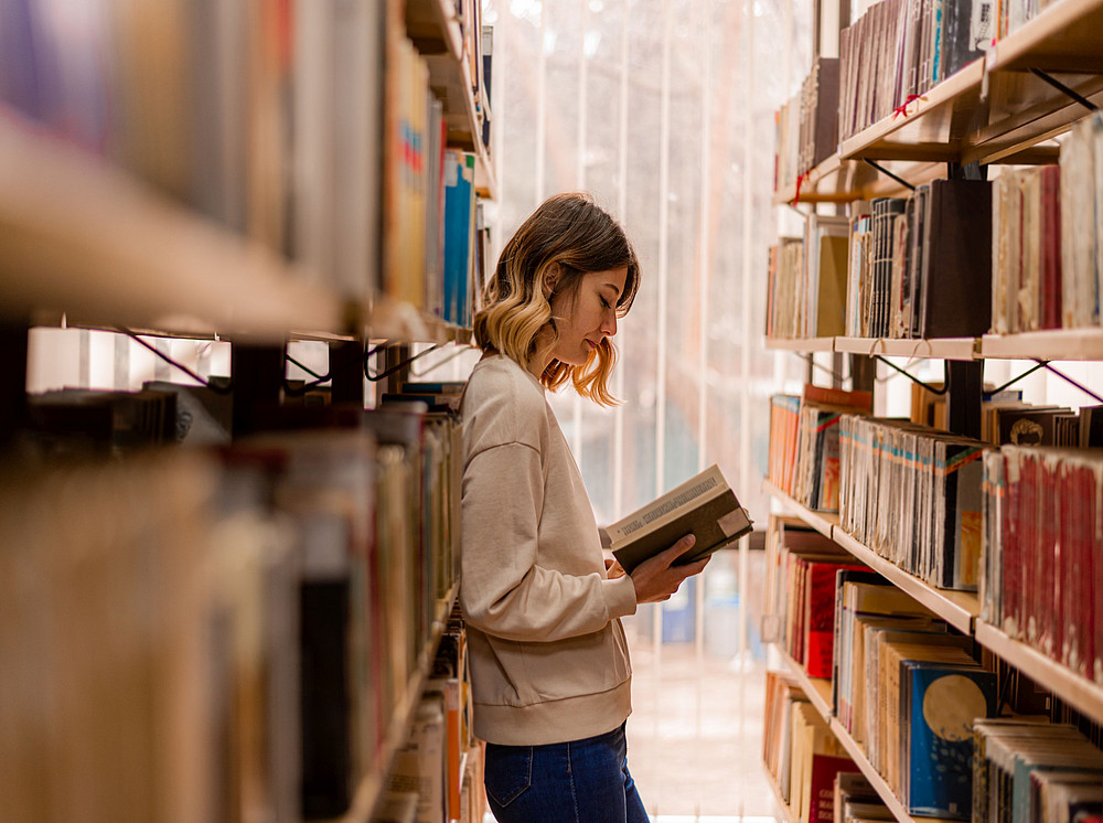 Person steht lesend zwischen zwei Regalen der Institutsbibliothek ©qunica.com - stock.adobe.com