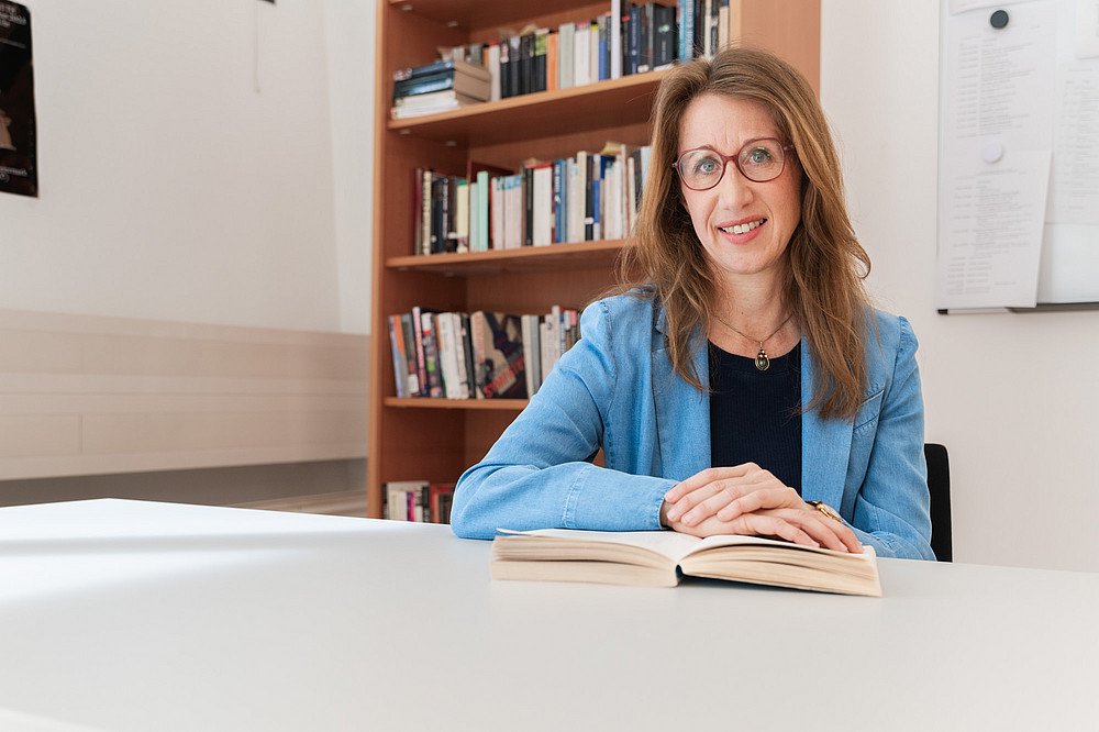 Portrait Julia Hoydis, sitzend an einem Tisch vor einem Bücherregal ©Uni Graz/Angele