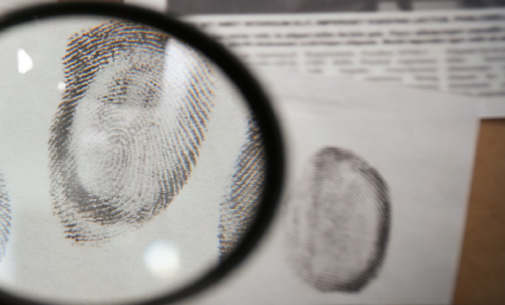 Eine Lupe vergrößert einen Fingerabdruck auf einem Blatt Papier, während im Hintergrund weitere Fingerabdrücke und ein unscharfer Zeitungsausschnitt zu sehen sind. Das Bild vermittelt den Eindruck einer kriminalistischen Untersuchung oder forensischen Analyse.
