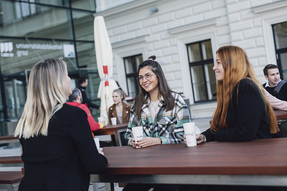 Studierende sitzen mit To-Go-Heißgetränken gemeinsam im Außenbereich an einem Tisch und unterhalten sich fröhlich.