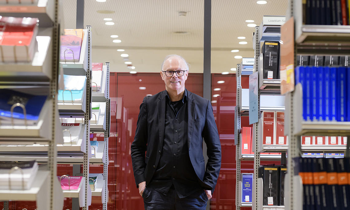 A person stands with their hands in their pockets between tall bookshelves in a brightly lit library. To the left and right, neat rows of books and magazines with different colors and covers are visible. In the background are red partitions and a ceiling with many round ceiling spotlights. The person is wearing dark, formal clothing and is looking straight ahead.