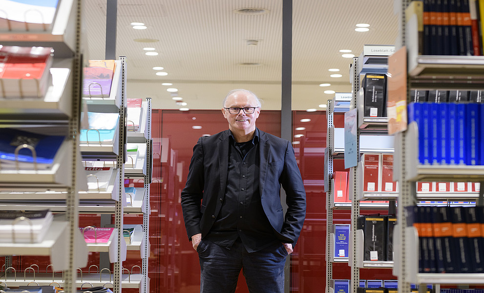 Eine Person steht mit Händen in den Taschen zwischen hohen Bücherregalen in einer hell beleuchteten Bibliothek. Links und rechts sind geordnete Reihen von Büchern und Zeitschriften mit verschiedenen Farben und Einbänden sichtbar. Im Hintergrund befinden sich rote Trennwände und eine Decke mit vielen runden Deckenspots. Die Person trägt dunkle, formelle Kleidung und blickt nach vorn.