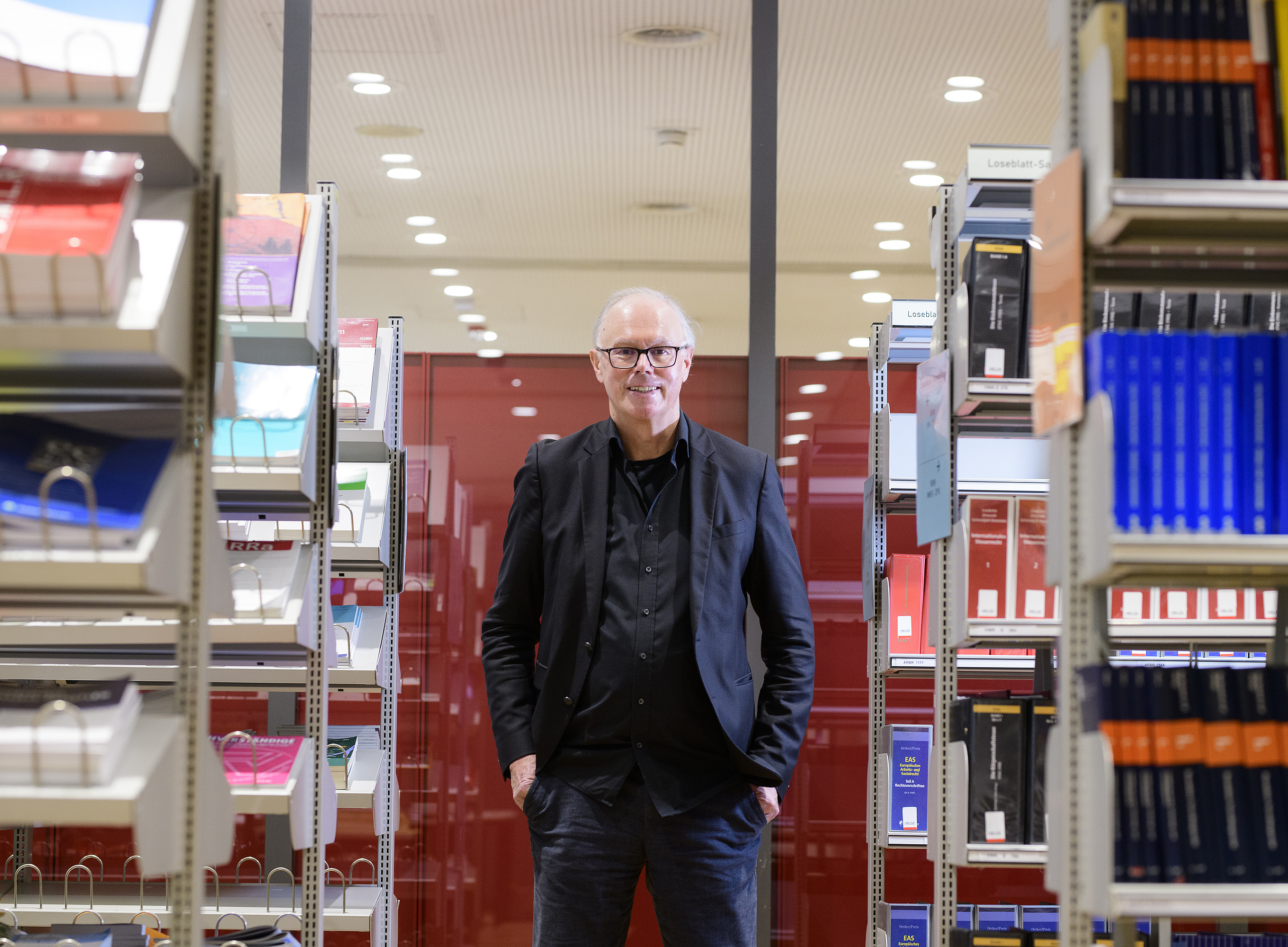 A person stands with their hands in their pockets between tall bookshelves in a brightly lit library. To the left and right, neat rows of books and magazines with different colors and covers are visible. In the background are red partitions and a ceiling with many round ceiling spotlights. The person is wearing dark, formal clothing and is looking straight ahead. ©Uni Graz/Tzivanopoulos