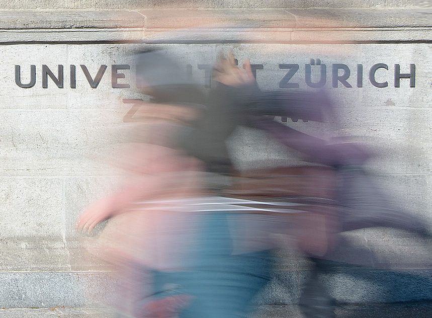 Schriftzug Universität Zürich an grauer Mauer hinter einer verschwommenen Person in Bewegung ©UZH Kommunikation