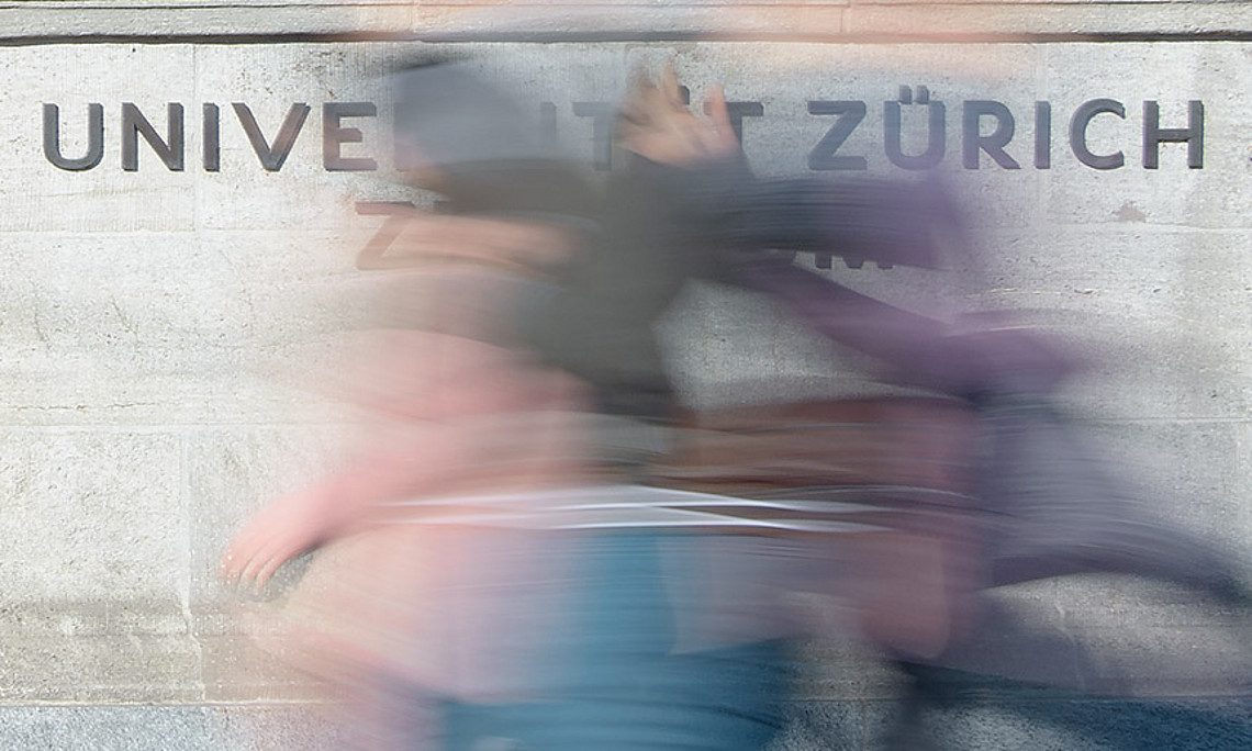 Schriftzug Universität Zürich an grauer Mauer hinter einer verschwommenen Person in Bewegung