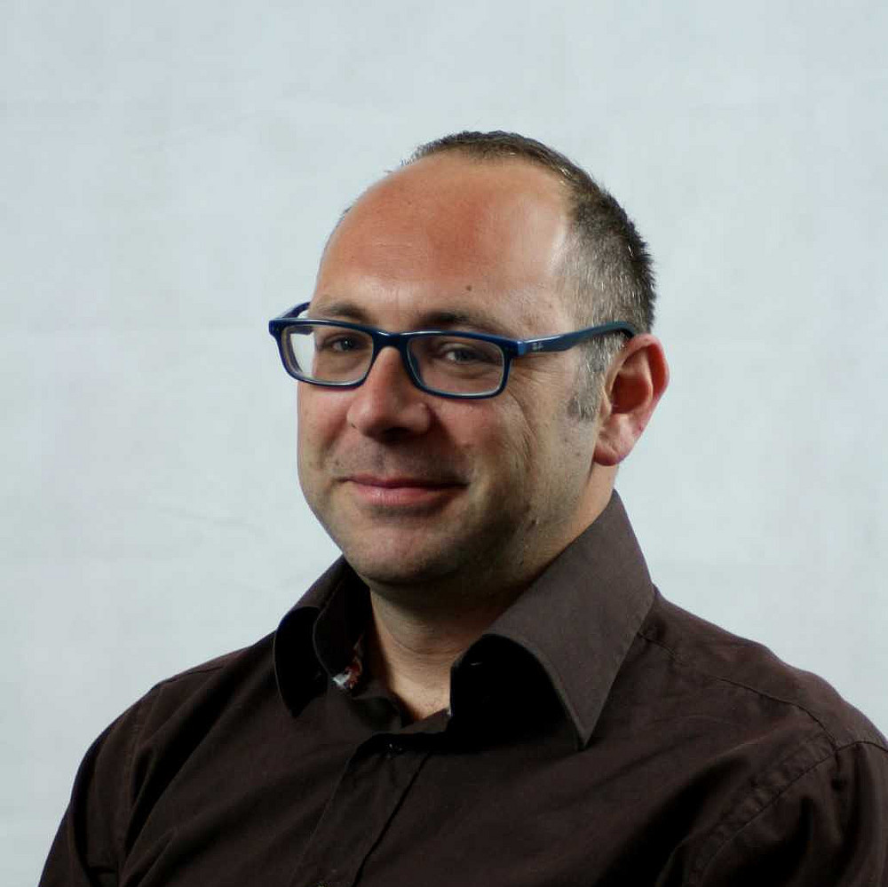 Porträt von Ben Gidley, einem Mann mit kurz geschnittenem Haar und hoher Stirn, der eine blaue Brille und ein dunkles Hemd trägt. Er blickt leicht seitlich in die Kamera und lächelt dezent vor einem hellen, neutralen Hintergrund. ©Ben Gidley